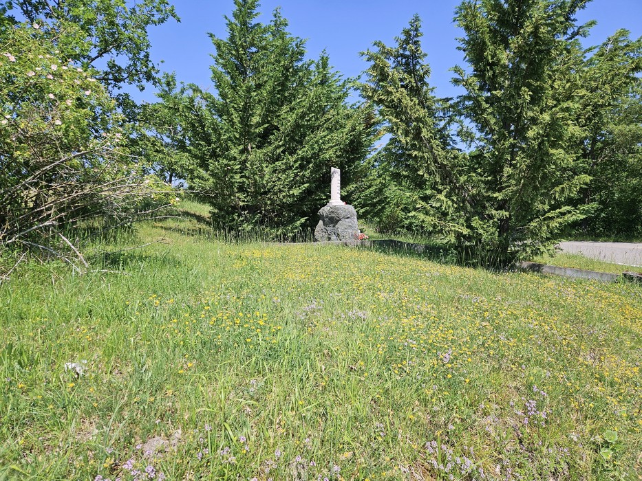 Cippo in pietra con iscrizione su un prato, circondato da alberi e cespugli lungo una strada.