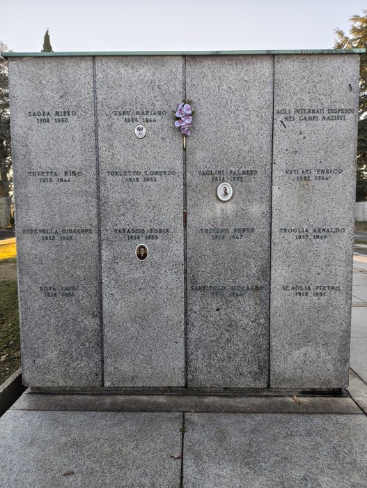 Monumento commemorativo con lapidi e fiori, situato nel Campo della Gloria, circondato da alberi.