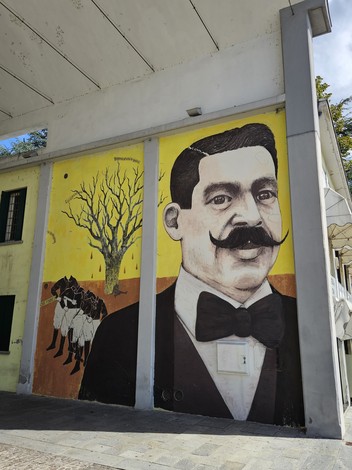 Murale colorato che raffigura un uomo con baffi e papillon, sullo sfondo un albero secco e cavalli.