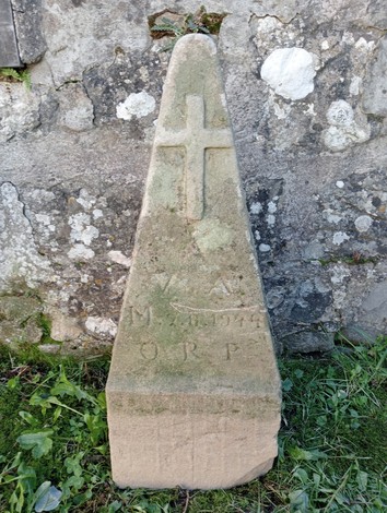 Cippo commemorativo di Videschi, con superficie ruvida e decorato da una croce, immerso nella vegetazione.