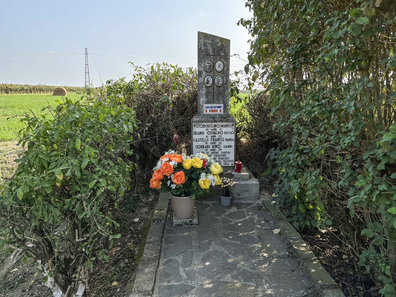 Cippo commemorativo decorato con fiori, situato in un ambiente rurale.