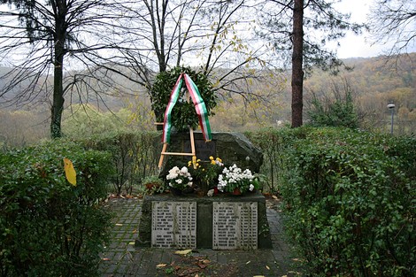 Monumento ai caduti di Polcanto, circondato da fiori e piante, in un contesto naturale sereno.