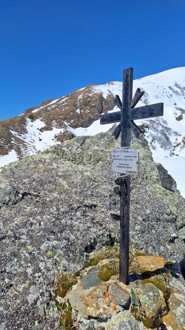 Croce metallica su una roccia coperta di muschio, con sfondo innevato e cielo blu chiaro.