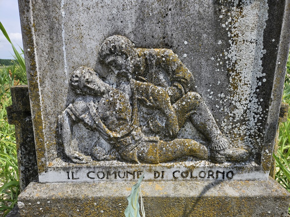 Cippo commemorativo con scultura di due figure, immerso in un paesaggio naturale e circondato da erba alta.