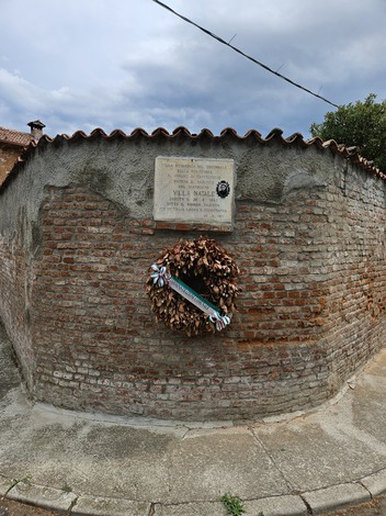 Lapide commemorativa su un muro di mattoni con una corona di foglie secche e nastri colorati.