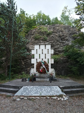 Monumento ai caduti di Rio Farnese, con una corona di fiori e un contesto naturale di rocce e vegetazione.