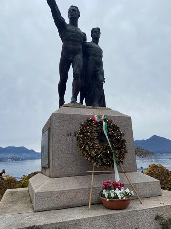 Monumento ai caduti con statue di soldati e una corona d'alloro ai piedi, vista lacustre sullo sfondo.