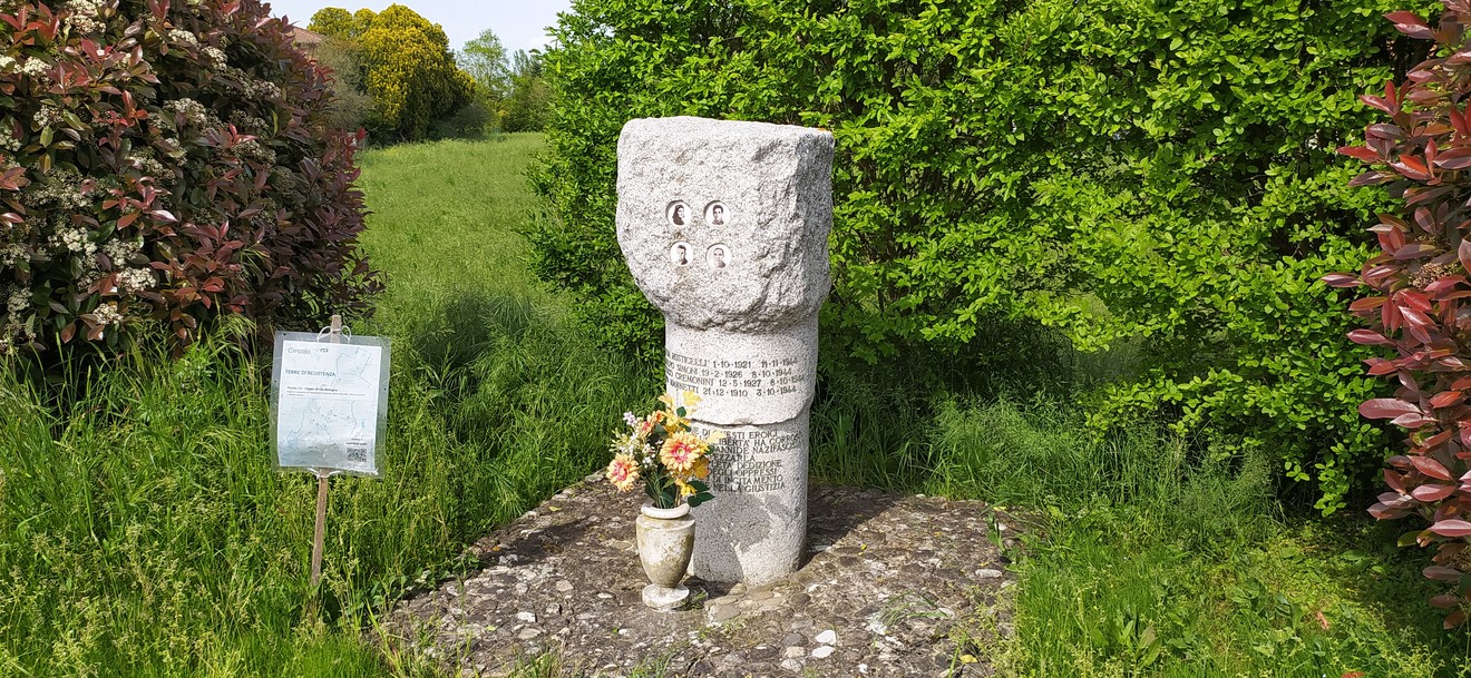 Cippo commemorativo circondato da vegetazione, con fiori e un cartello informativo nelle vicinanze.