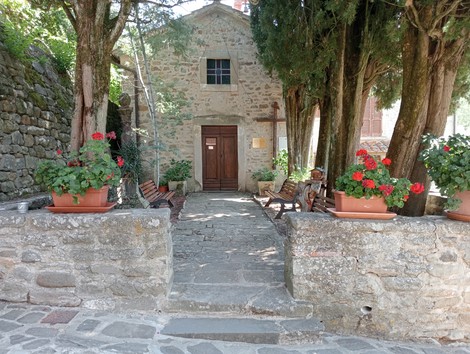 Facciata di una storica struttura in pietra circondata da alberi e fiori rossi, con panchine e un'atmosfera accogliente.