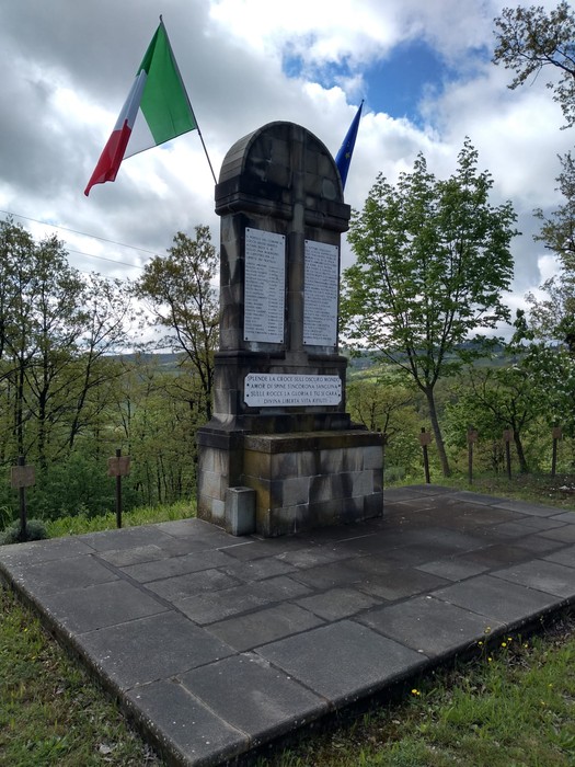 Monumento ai martiri dei Boschi di Ciano, circondato da alberi e bandiere italiane ed europee.