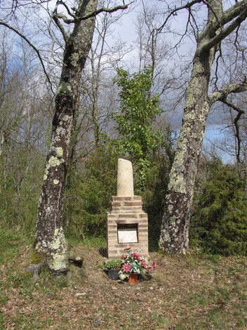 Cippo commemorativo incastonato tra due alberi, circondato da fiori in un ambiente naturale tranquillo.