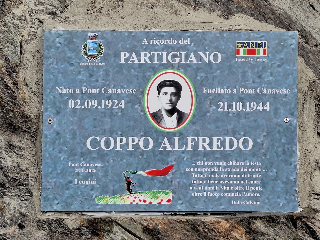 Targa commemorativa in metallo su parete rocciosa, con ritratto del partigiano e iscrizione.