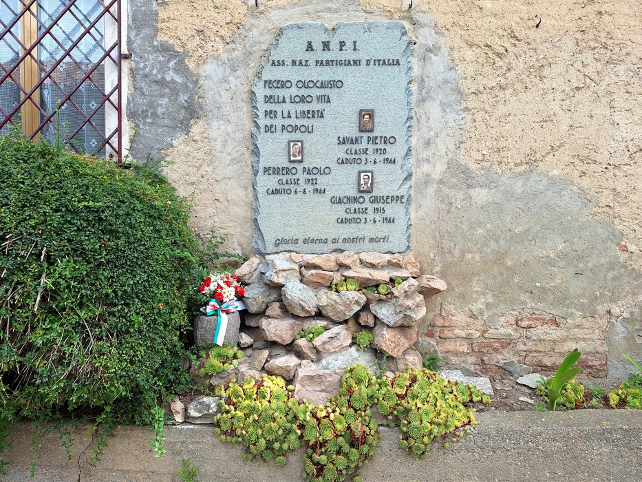 Lapide commemorativa in pietra con nomi di partigiani e decorazioni floreali, collocata su un muro roccioso.