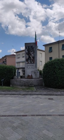 Monumento ai partigiani con bandiera italiana, situato in un'area cittadina recintata da siepi verdi.