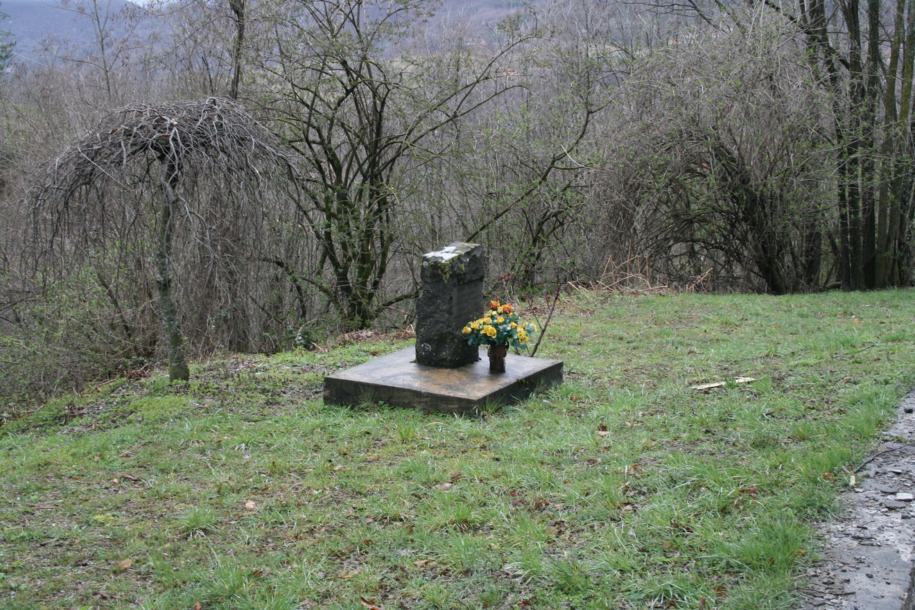 Cippo commemorativo su un prato, circondato da alberi e vegetazione, con fiori posati davanti.