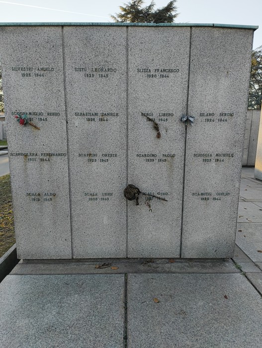 Monumento commemorativo al Campo della Gloria con iscrizioni e omaggi floreali. Atmosfera di rispetto e memoria.