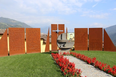 Monumento a Seghezzi con pannelli di metallo e fiori rossi, situato in un paesaggio montano.