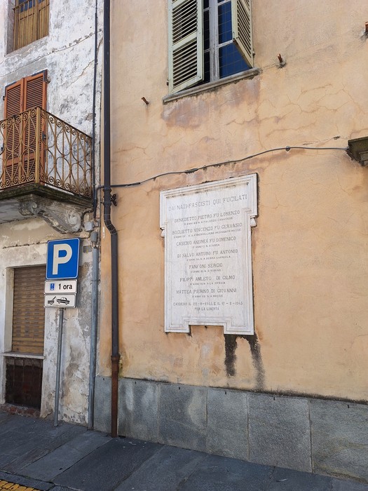 Lapide dedicata ai VII Martiri su una parete di un edificio, con un parcheggio limitrofo.