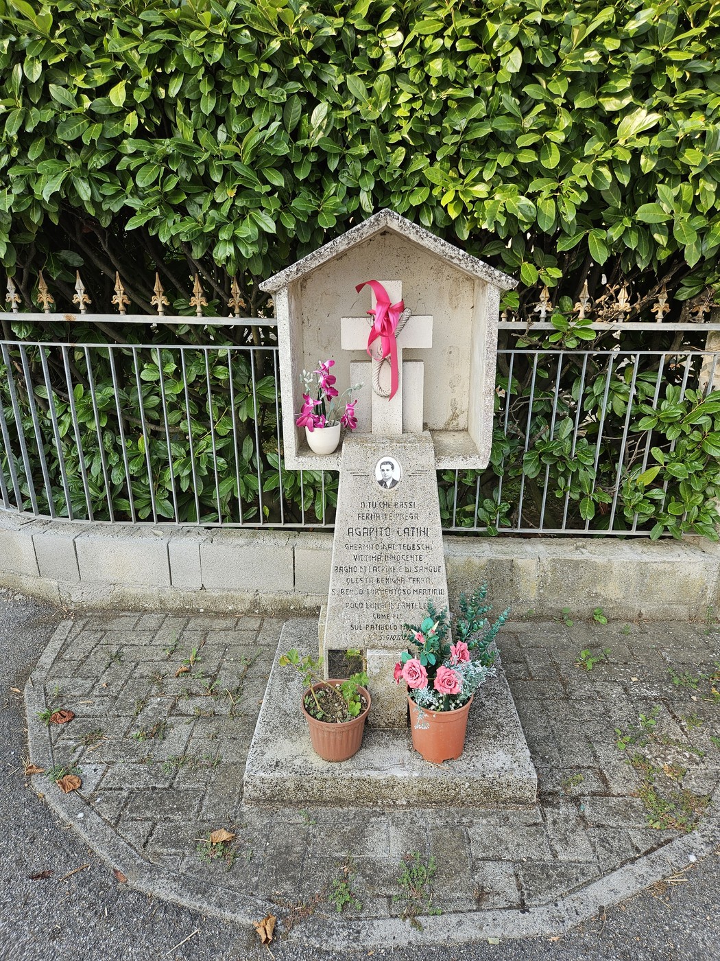 Cippo commemorativo con fiori e decorazioni, dedicato ad Agapito Latini, circondato da vegetazione.