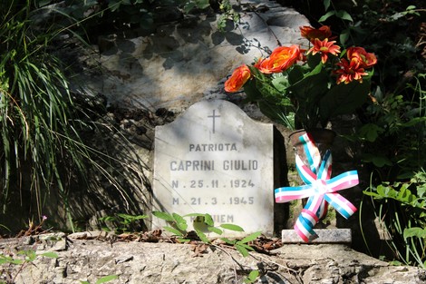 Lapide commemorativa di Caprini Giulio, con fiori e un nastro colorato, immersa nella vegetazione.