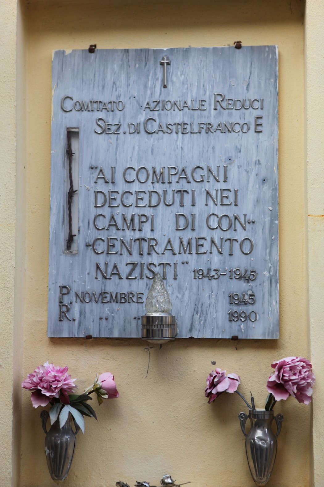 Lapide commemorativa dedicata ai caduti nei campi di concentramento nazisti, con fiori artificiali ai lati.