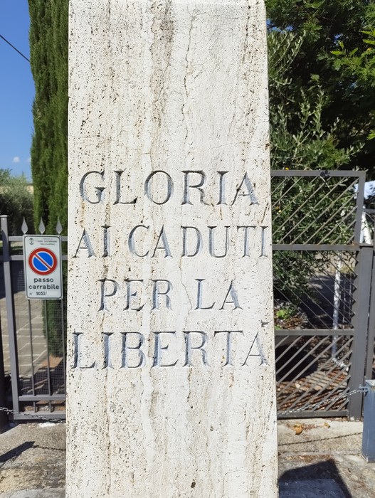 Cippo in pietra con iscrizione, posto all'aperto davanti a un cancello in ferro.