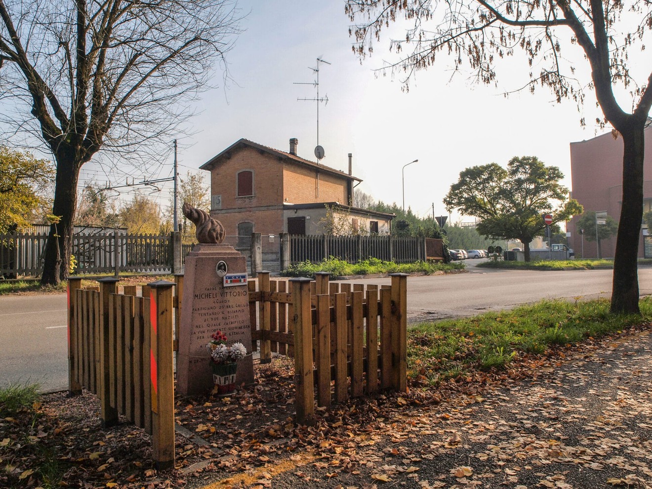 Cippo commemorativo dedicato a Micheli, circondato da alberi e recinzione, vicino a una strada.