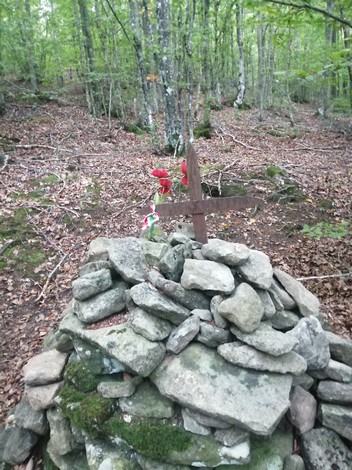 Cippo commemorativo circondato da alberi, con una croce di legno e adornato da fiori nel bosco.
