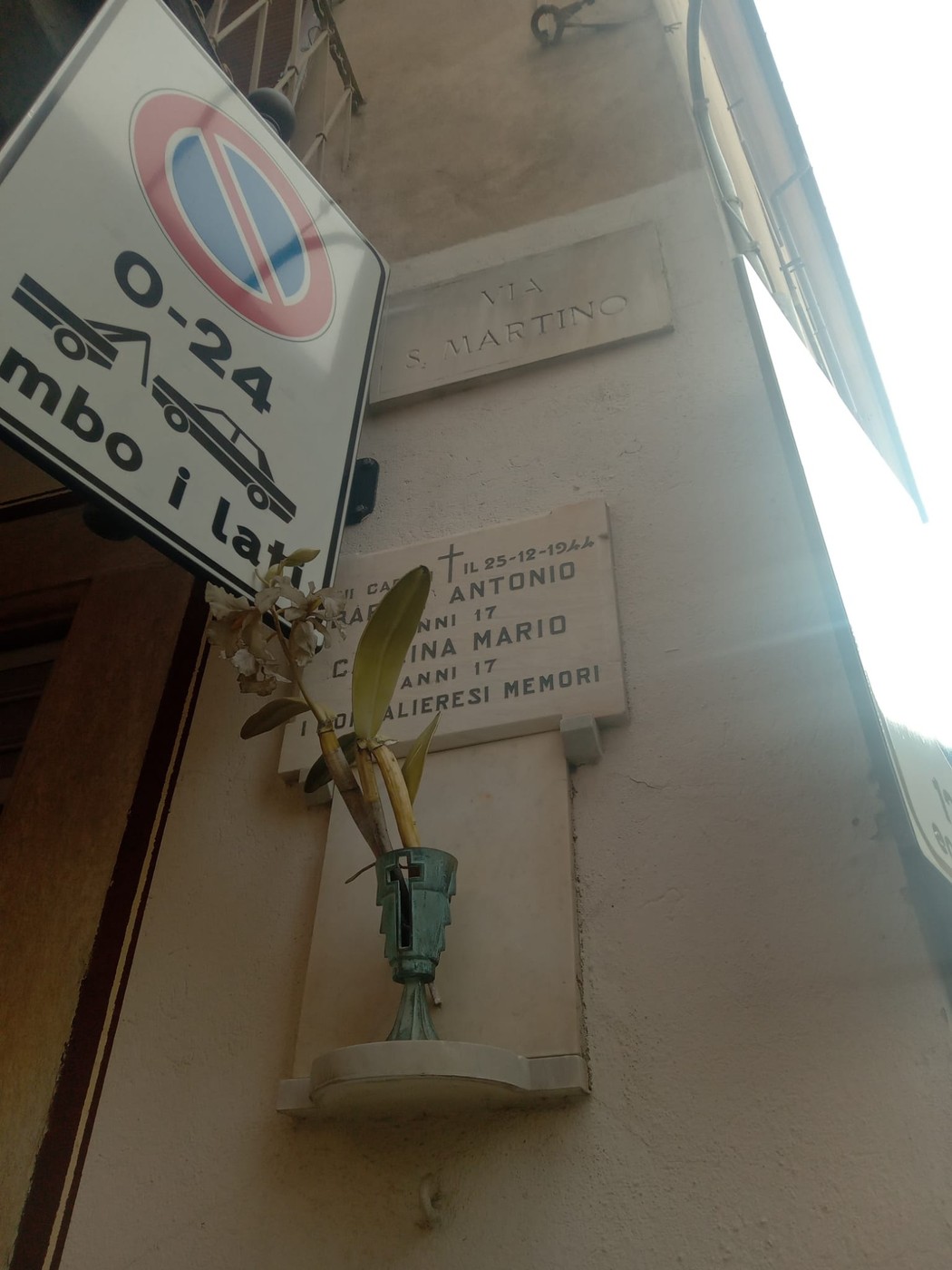 Lapide commemorativa in una parete, con fiori e segnaletica stradale visibile.
