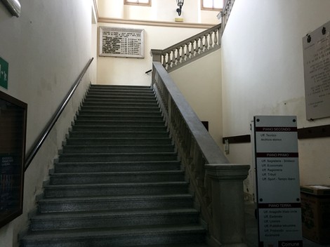 Scalinata all'interno di un edificio, con una lapide commemorativa sulla parete e segnaletica direzionale visibile.