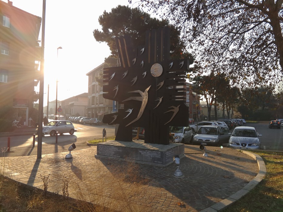 Monumento ai Caduti in metallo scuro su una rotatoria, con sagome di uccelli e iscrizione alla base.