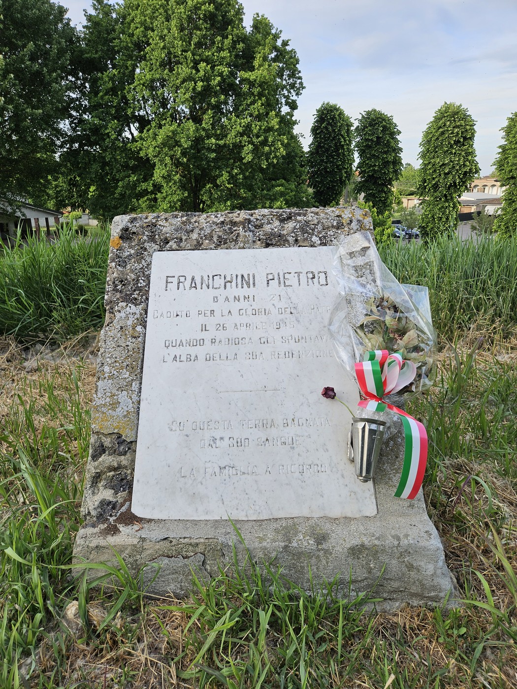 Cippo commemorativo a Franchini Pietro, con fiori e nastri tricolori, immerso nel verde di un campo.