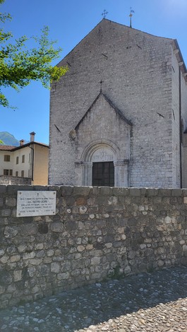 Lapide commemorativa vicino alla chiesa in pietra, sullo sfondo un cielo blu e alberi verdi.