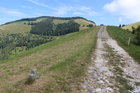 Percorso in un paesaggio collinare, con una croce visibile lungo il sentiero e verdeggianti montagne sullo sfondo.
