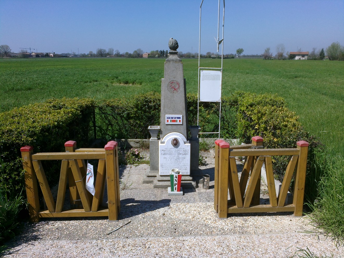Cippo commemorativo a Campedelli, circondato da un prato verde, con ornamenti e elementi decorativi.