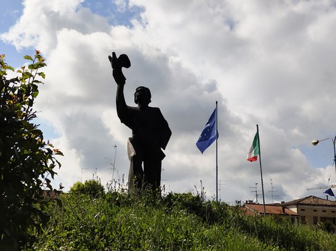 Monumento al Partigiano con figura che saluta, circondato da bandiere e un cielo nuvoloso.