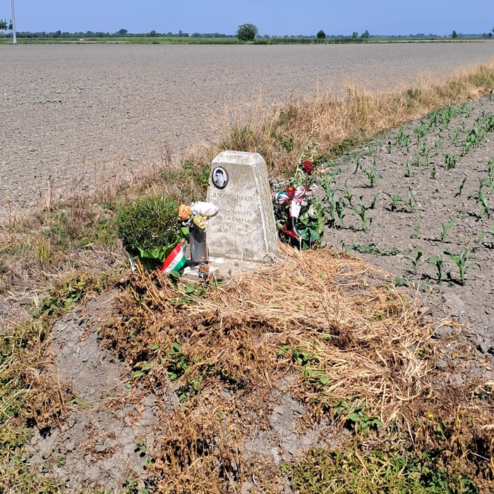 Cippo a Lanza con fiori e bandiere, circondato da un campo agricolo in un paesaggio rurale.