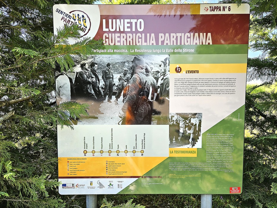 Pannello informativo "Tappa n. 6" con foto storiche e testi sulla Resistenza, tra alberi di conifere.
