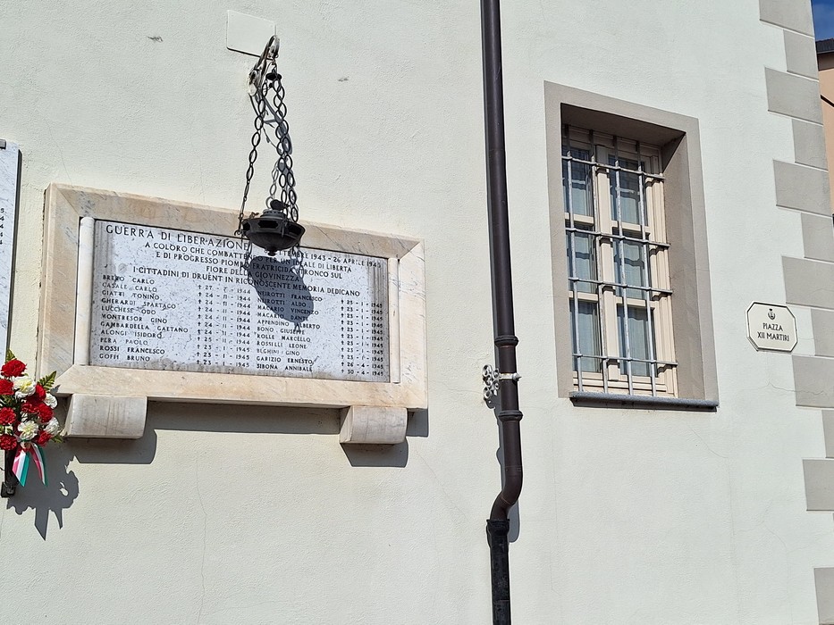 Lapide in marmo con iscrizione, appesa alla parete di un edificio, con lanterna e mazzo di fiori accanto.