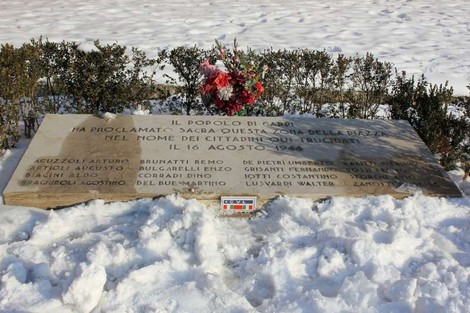 Monumento dedicato ai martiri del 16 agosto, con fiori e pietra commemorativa in un paesaggio innevato.