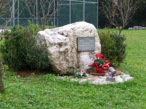 Lapide commemorativa su roccia con fiori e decorazioni in un contesto verde e naturale.