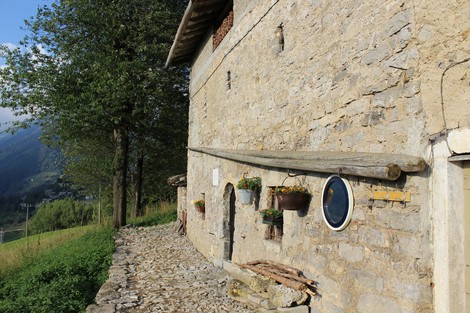 Edificio rustico in pietra con finestre circolari e fiori, circondato da verde e montagne.