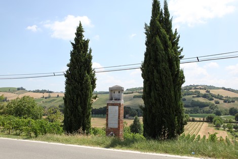 Cippo commemorativo tra due cipressi, affacciato su un paesaggio collinare e agricolo.