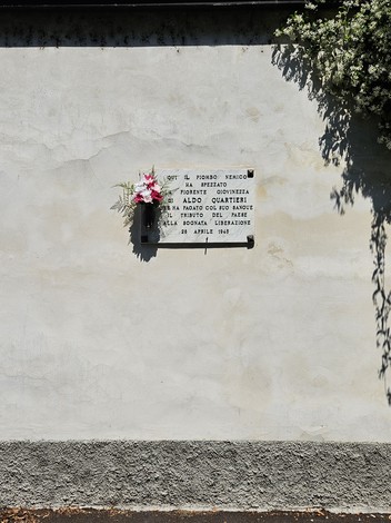 Lapide commemorativa su una parete con fiori, in memoria di Aldo Quartieri e altri.