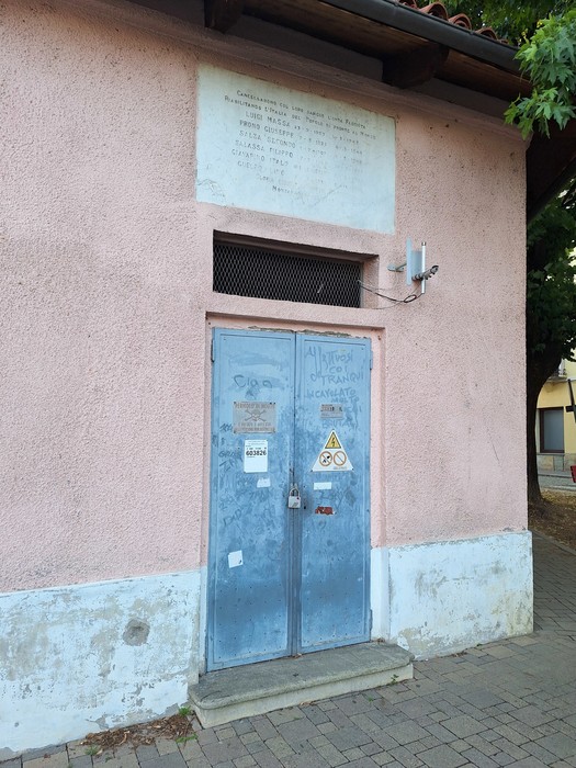 Lapide con iscrizione sopra una porta blindata grigia, sulla facciata rosa di un edificio.