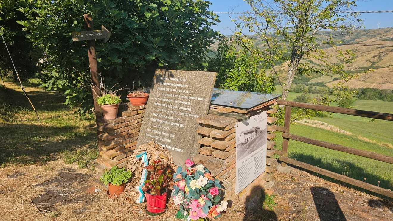 Monumento per Ca' di Guzzo, circondato da piante e fiori, con vista su un paesaggio collinare.