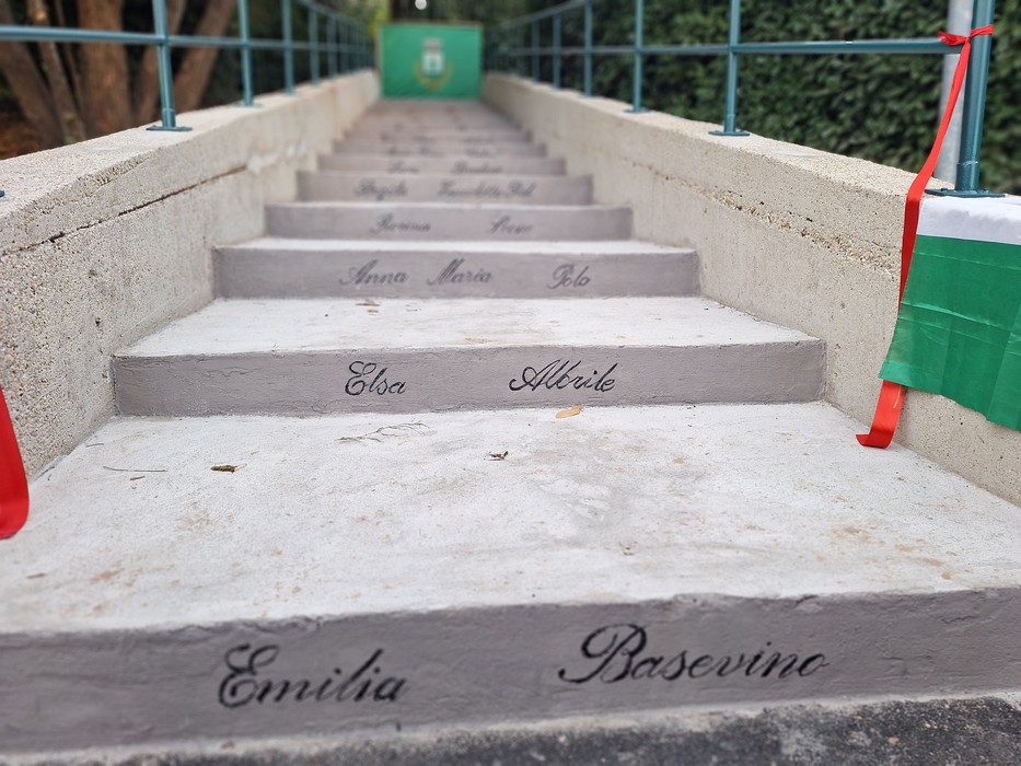 Scalinata in pietra con nomi di donne incisi su ogni gradino, fiancheggiata da ringhiere e bandiere tricolori.