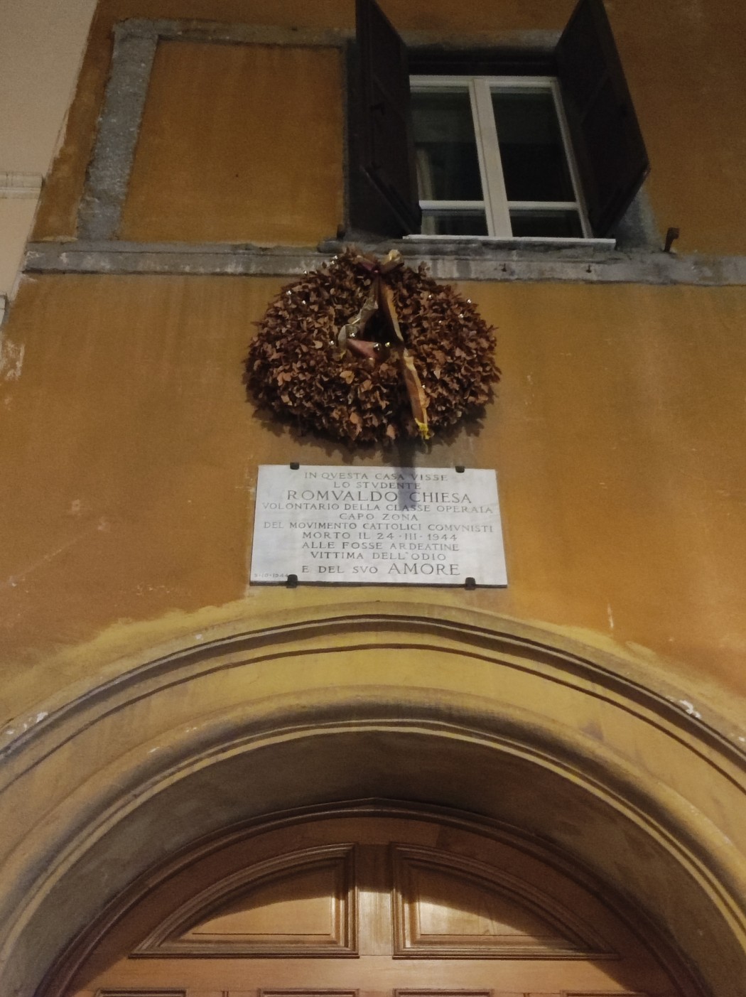 Lapide commemorativa sulla facciata di una chiesa, adornata con una corona di foglie secche.