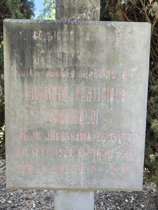 Lapide in pietra con iscrizione dedicata alla divisione partigiana Garibaldi, tra la vegetazione.