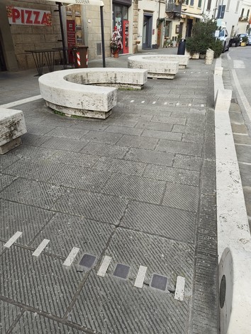 Pietre d'inciampo sulla pavimentazione di una strada urbana, circondate da panchine in marmo.